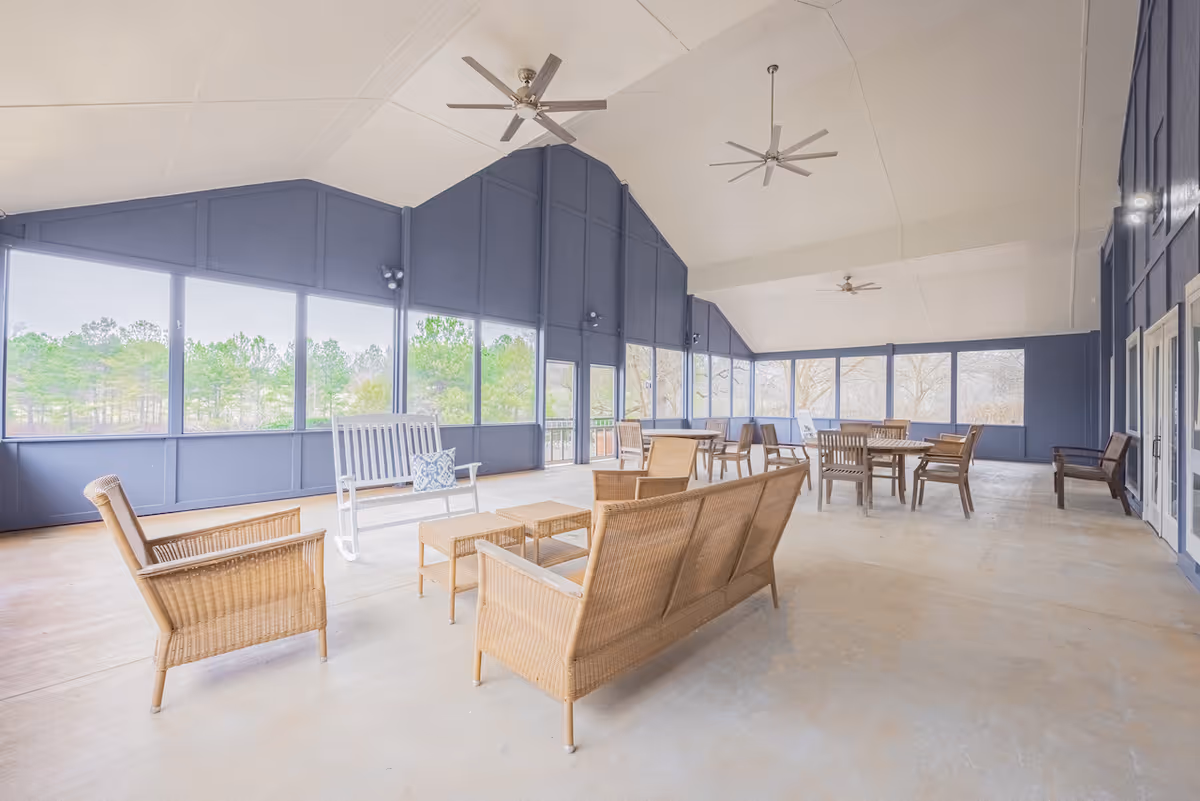 A spacious screened-in porch area with wicker seating including chairs, a loveseat, and a white rocking bench with a decorative pillow. Several wooden tables and chairs are arranged throughout the space. The porch has a high ceiling with three ceiling fans and large screened windows offering a view of trees outside.