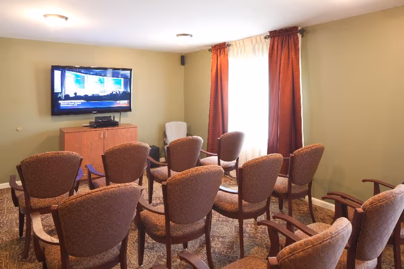 A small room with multiple upholstered chairs arranged in rows facing a wall-mounted flat screen TV. The room has beige walls, a patterned carpet, a wooden cabinet under the TV, and a window with sheer white curtains and red drapes.