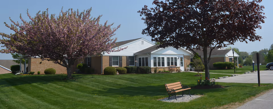 Single-story residential care building with a manicured lawn, flowering trees, and a bench near the entrance.