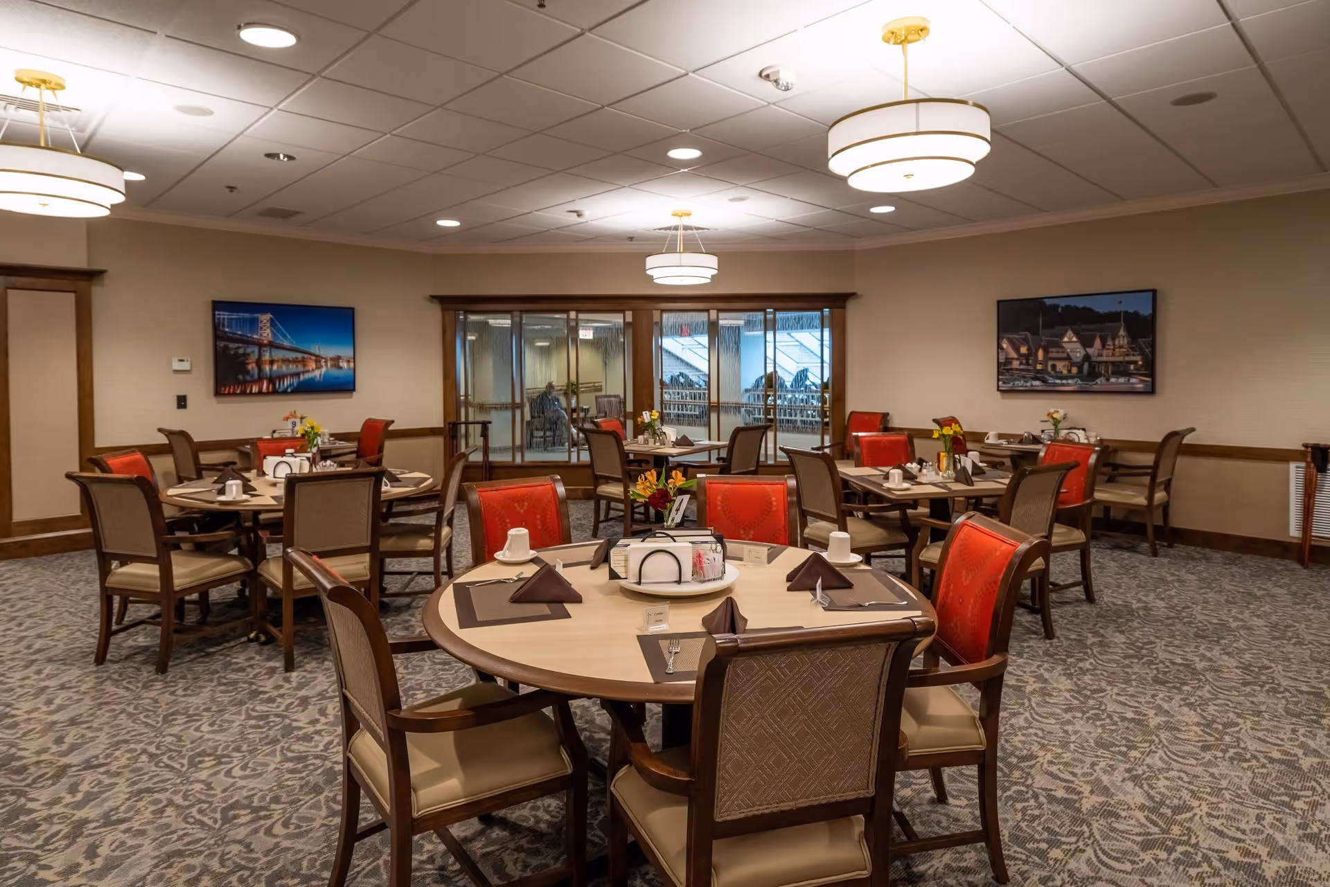 A dining room in a senior living facility with round tables set for meals. Each table has placemats, folded napkins, cups, and small flower arrangements. The room has carpeted floors, beige walls, and framed pictures on the walls. There are large glass doors at the back of the room.