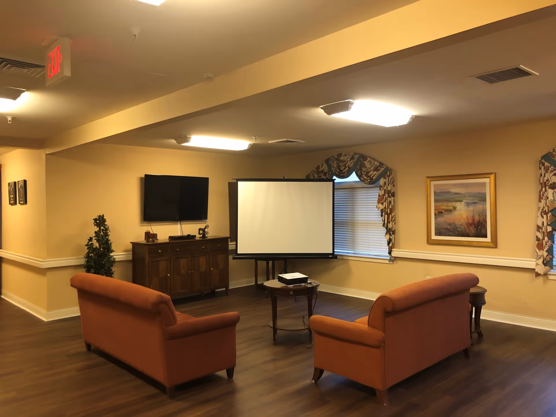 A cozy living room area with two orange sofas facing a TV mounted on the wall and a projector screen. The room has wooden flooring, beige walls, a framed painting, and windows with patterned curtains. There is a small round table with a projector on it between the sofas.