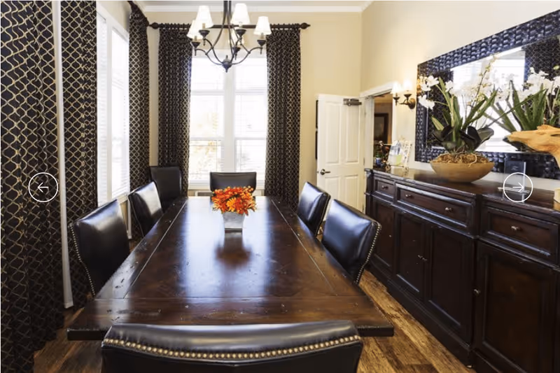 A dining room with a long dark wooden table surrounded by six black leather chairs. The table has a small floral centerpiece with orange and red flowers. The room features large windows with patterned dark curtains, a chandelier hanging above the table, and a dark wooden sideboard with decorative plants and a large mirror on the wall.