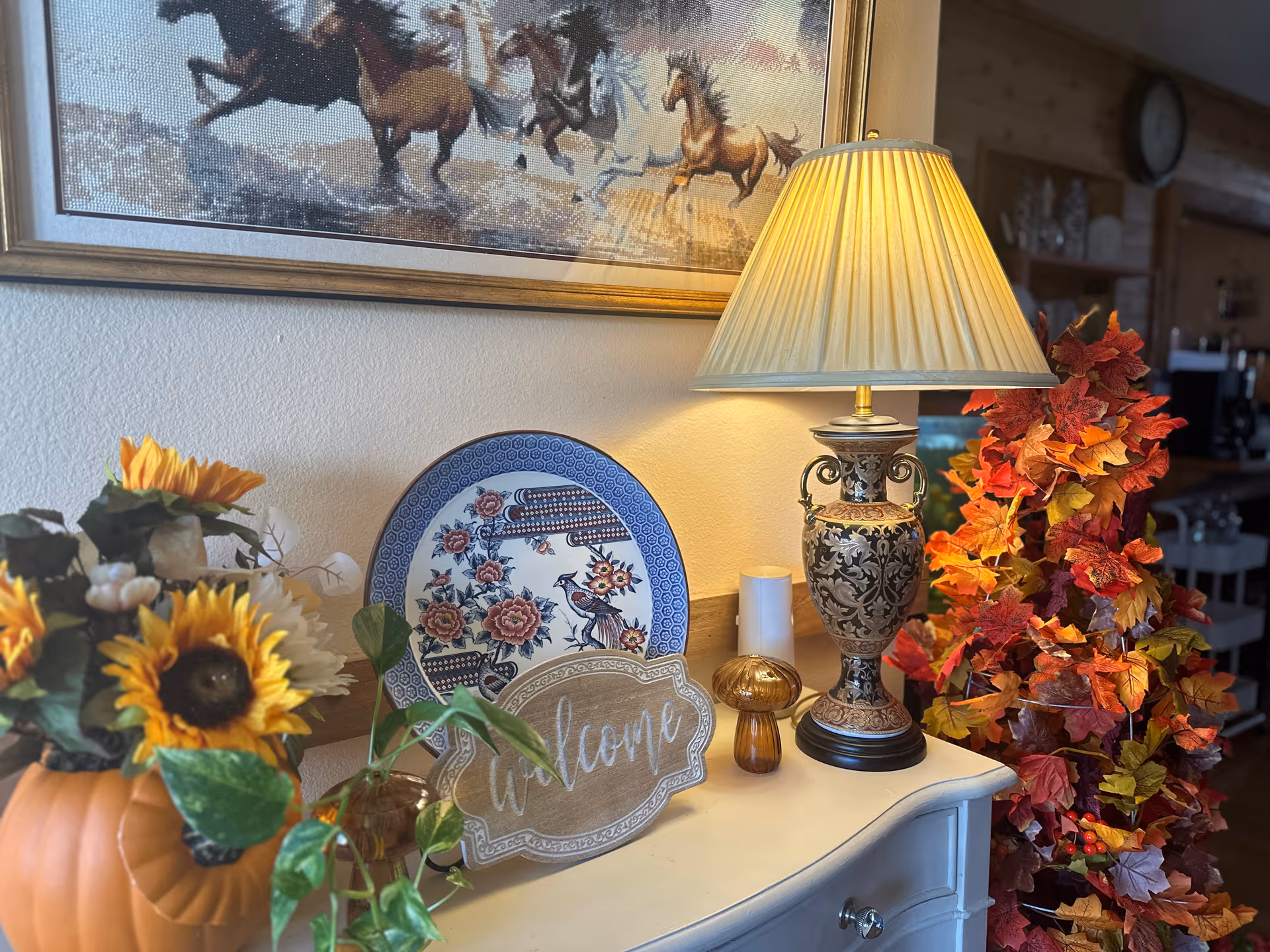 A decorative corner featuring a table with a vintage-style lamp, a blue and white floral plate, a wooden welcome sign, a small amber mushroom-shaped ornament, and a white candle. To the left, there is a pumpkin-shaped vase with sunflowers and other flowers. To the right, there is a tall arrangement of autumn-colored leaves. Above the table is a framed picture of running horses. The background shows a cozy interior with wooden walls and shelves.