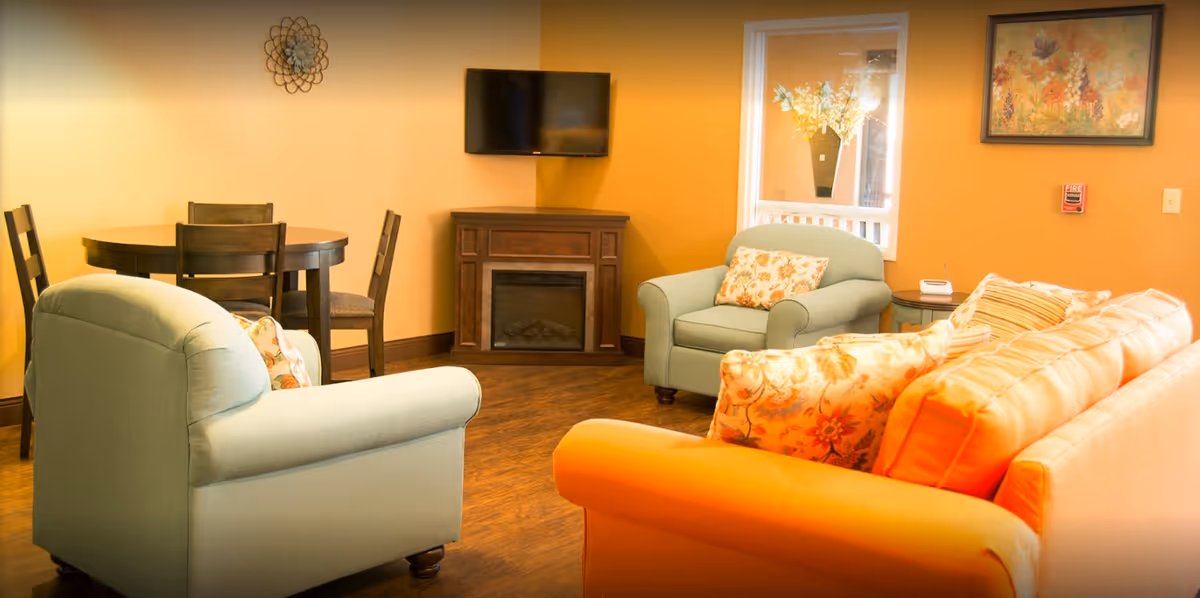 A cozy living room area with a light green armchair, an orange sofa with floral cushions, a small round wooden dining table with four chairs, a wall-mounted TV above a wooden electric fireplace, and a window with a vase of flowers. The walls are painted warm yellow and decorated with a floral painting and a decorative metal wall piece.