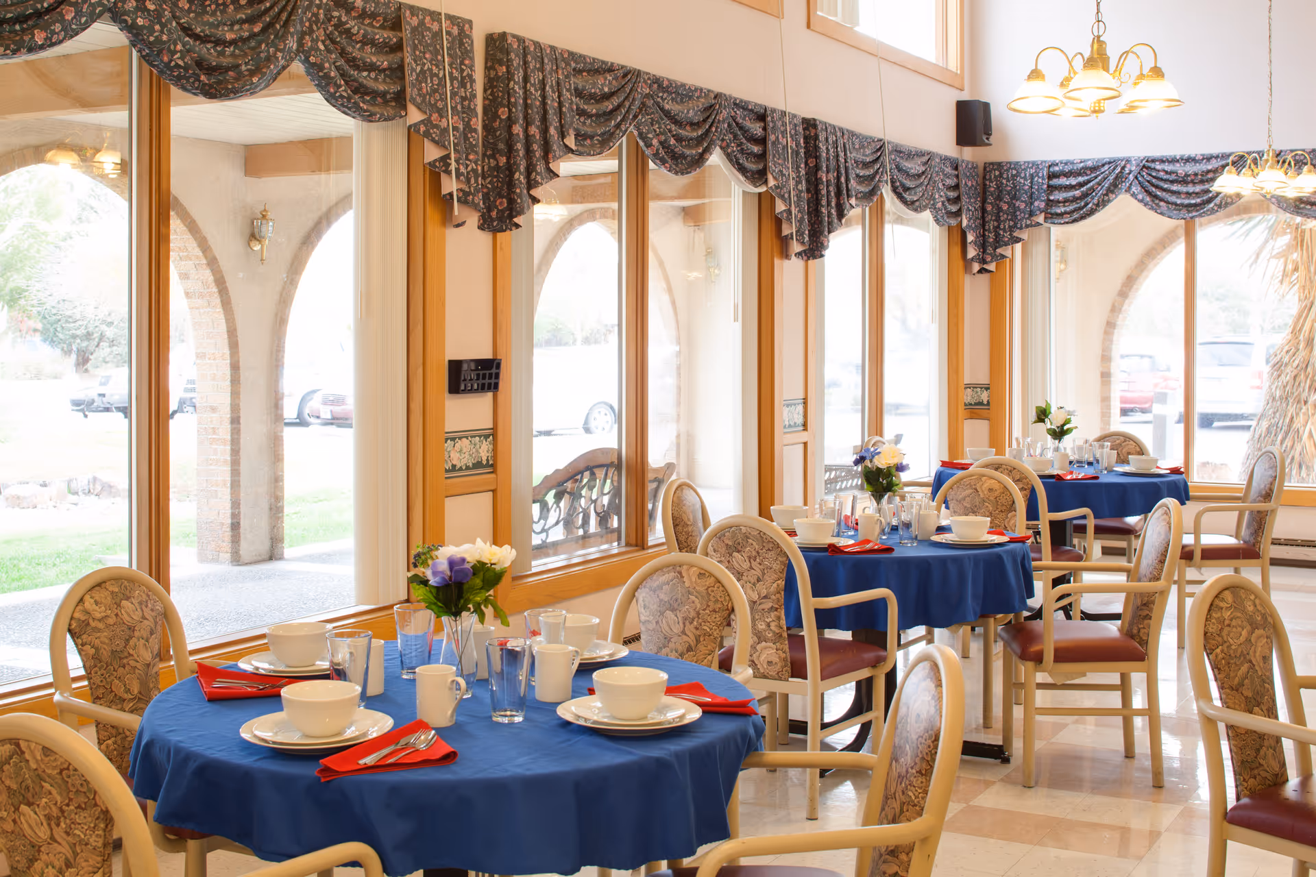 Sunlit dining room with round tables covered in blue tablecloths, place settings and floral centerpieces beside large arched windows.