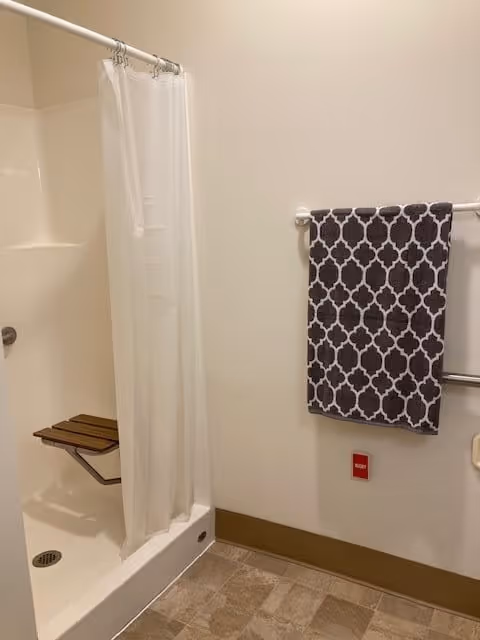 A bathroom with a white shower area featuring a foldable wooden seat and a white shower curtain. On the adjacent wall, there is a towel rack holding a dark towel with a white geometric pattern. Below the towel rack is a red emergency pull button. The floor has a tiled pattern.