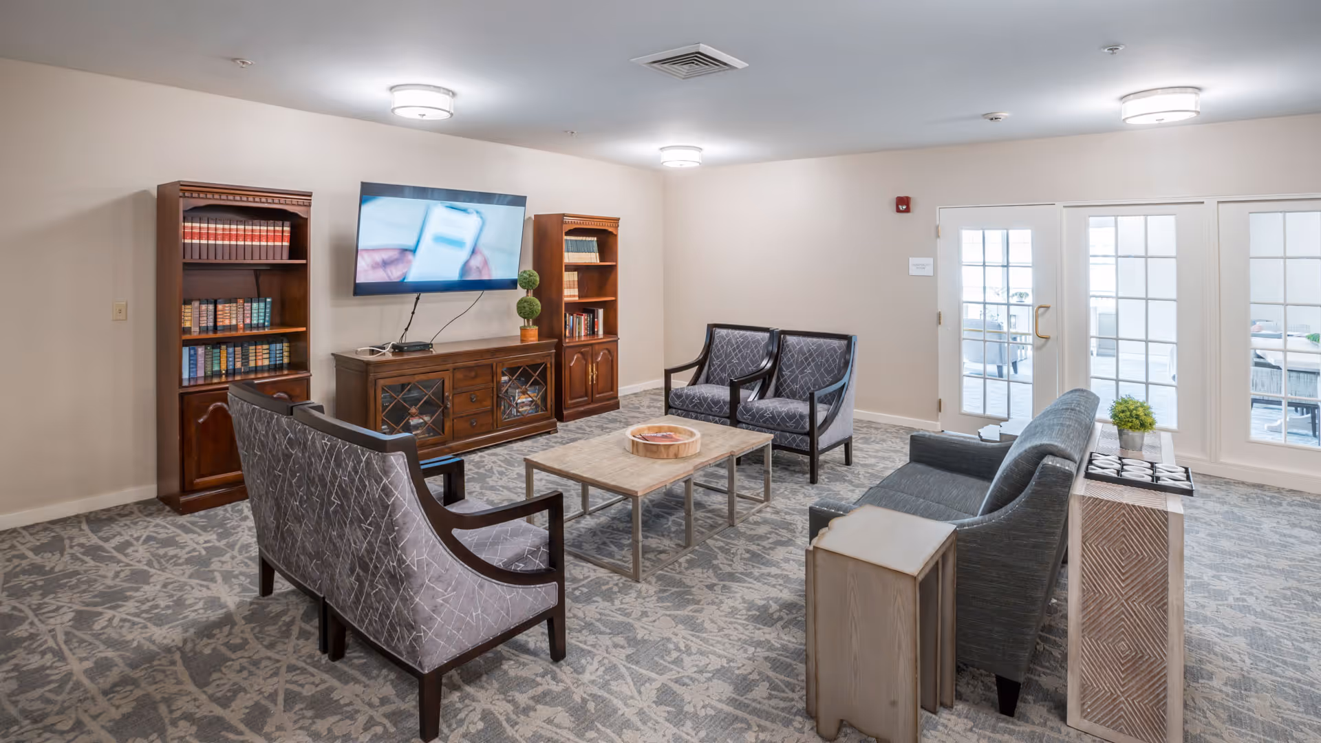 A well-lit living room area with patterned carpet, two wooden bookshelves filled with books, a flat-screen TV mounted on the wall, a wooden TV stand, a coffee table, and several upholstered chairs and sofas arranged around the table. French doors lead to an outdoor patio area.