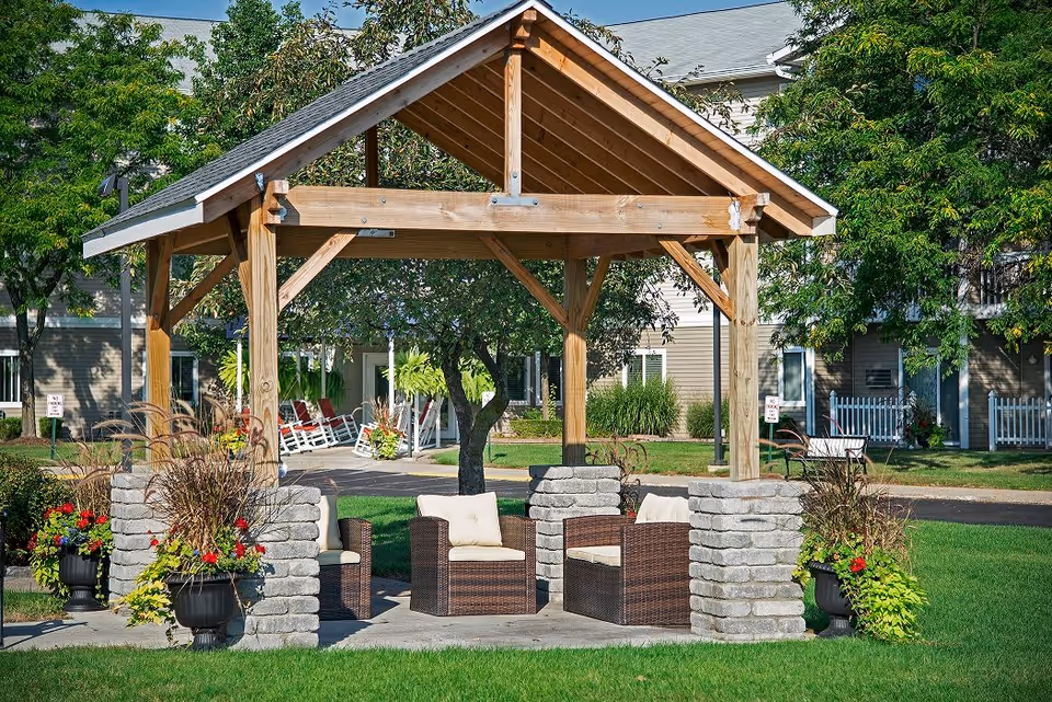 Outdoor seating area with a wooden pergola supported by stone pillars, surrounded by green grass, potted plants with flowers, and trees. In the background, there are buildings with windows and white picket fences.