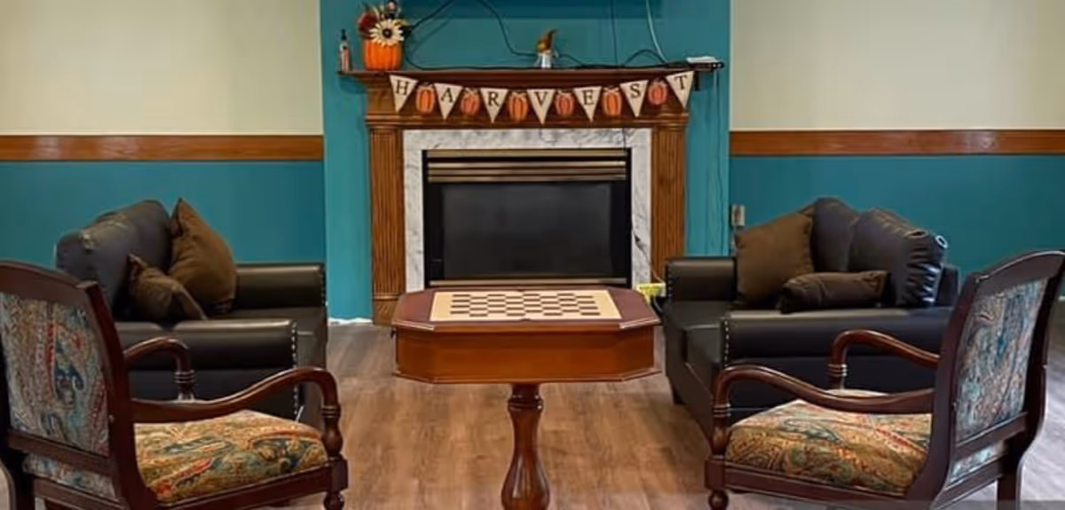 A cozy living room area with two dark leather sofas and two wooden armchairs with patterned cushions arranged around a wooden table with a checkerboard on top. In the background, there is a fireplace with a wooden mantel decorated with a 'HARVEST' banner and a small pumpkin decoration on the left side.