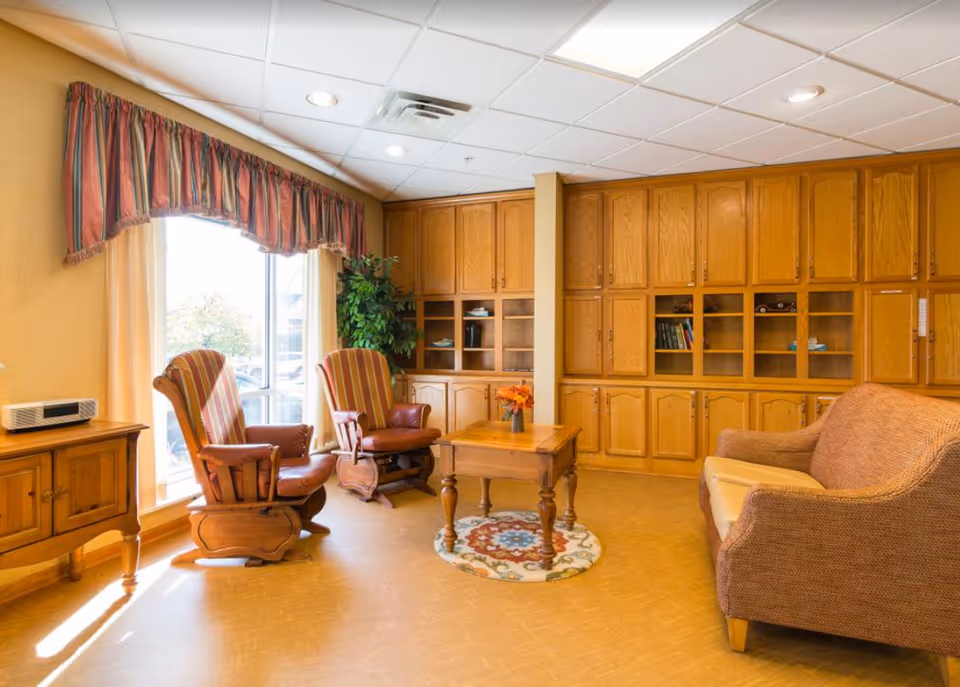 A cozy living room area with two wooden rocking chairs with striped cushions, a wooden coffee table with a small flower arrangement, a cushioned sofa, large wooden cabinets with shelves, a window with striped valance curtains, and a potted plant in the corner.