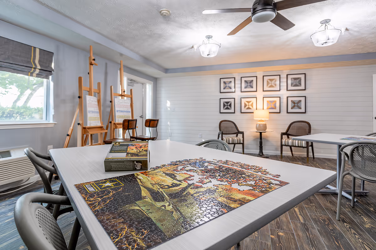 Well-lit activity room with a partially completed jigsaw puzzle on a table, easels and chairs, and framed artwork on the wall.