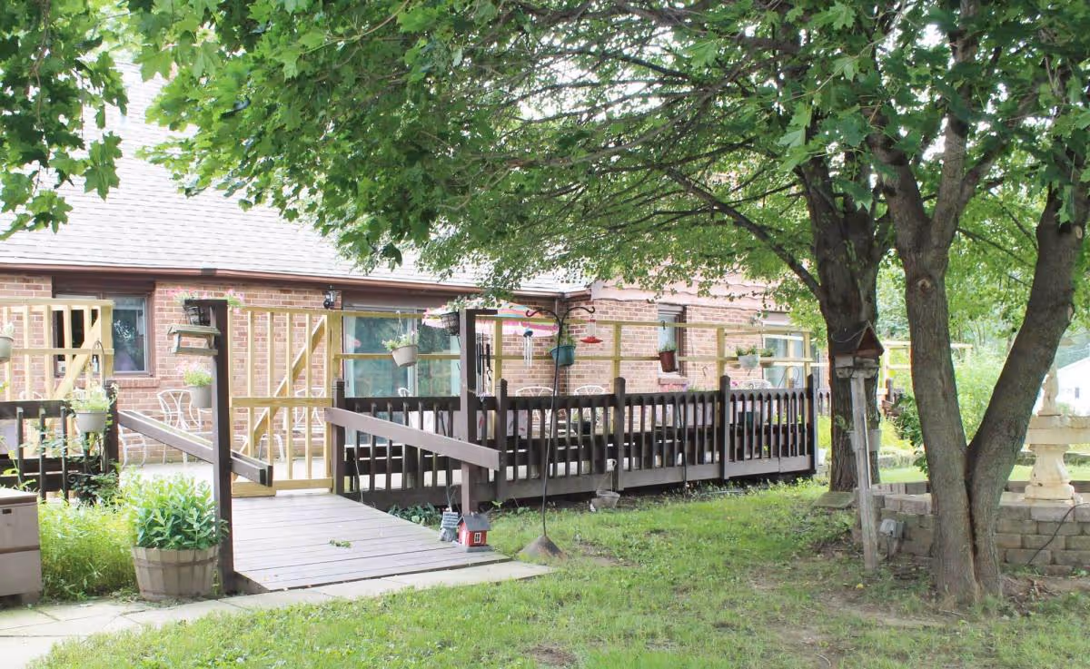 Outdoor view of a wooden deck attached to a brick building, surrounded by green grass and trees. The deck has a ramp and railing, with hanging flower pots and outdoor furniture visible. The area is shaded by large leafy trees.