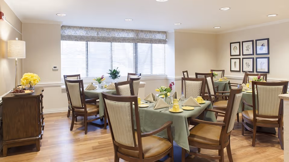 A bright dining room with several tables covered in green tablecloths, each set with yellow cups, plates, and beige napkins. Wooden chairs with cushioned seats surround the tables. A sideboard with a lamp and a vase of yellow flowers is against one wall, and framed botanical prints hang on another wall. Large windows with blinds allow natural light to fill the room.