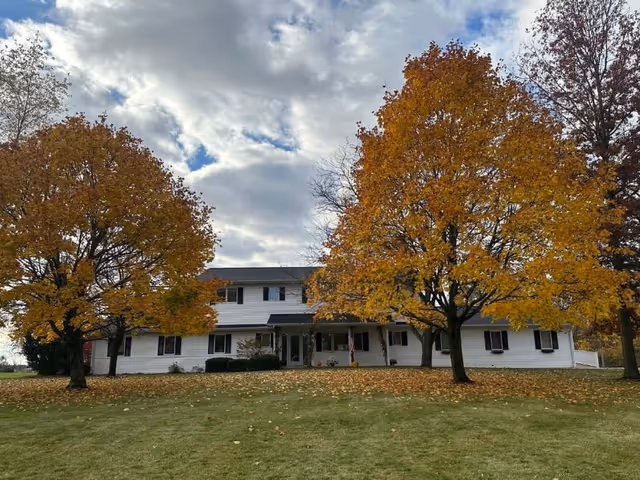 A large two-story white building with black shutters surrounded by trees with autumn foliage. The sky is partly cloudy and the ground is covered with fallen leaves and green grass.