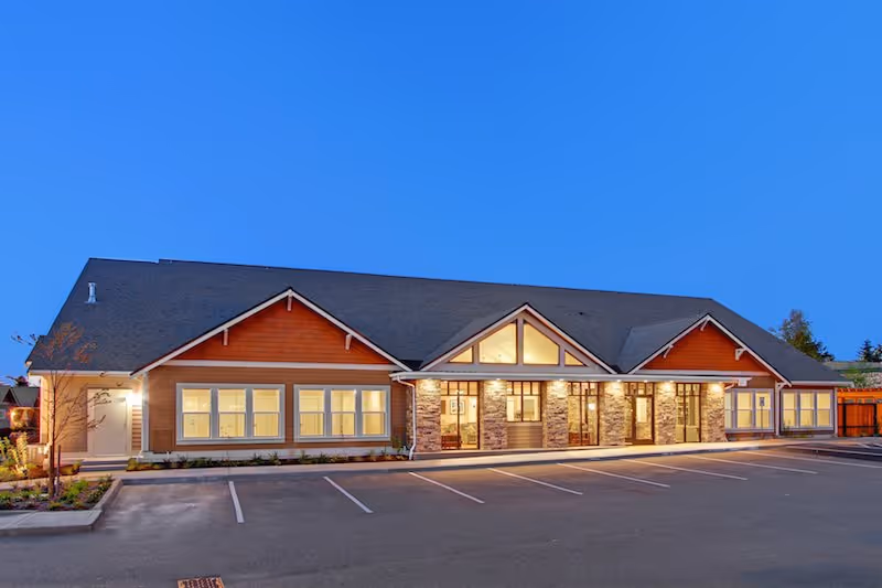 Exterior view of a single-story building at dusk with warm interior lights visible through multiple windows. The building has a pitched roof, stone pillars at the entrance, and a parking lot in front with marked parking spaces. The sky is clear and blue.