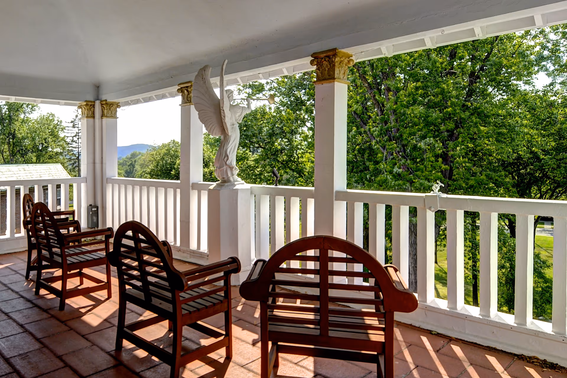A covered outdoor balcony with four wooden benches facing outward. The balcony has white railings and columns with gold decorative tops. A white angel statue blowing a trumpet is mounted on one of the railings. Green trees and a distant mountain are visible beyond the balcony.