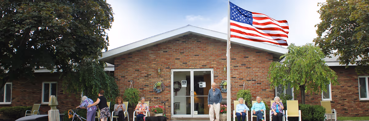 Front exterior view of a single-story brick building with a large American flag on a flagpole in front. Several elderly people are sitting and standing near the entrance, with some using walkers and others seated on chairs. There are trees and shrubs around the building under a clear blue sky.