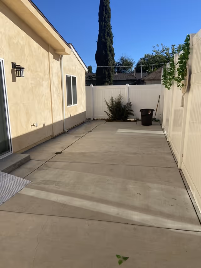 Concrete backyard patio between a beige house and white vinyl fence with a trash bin and a tall tree in the background.