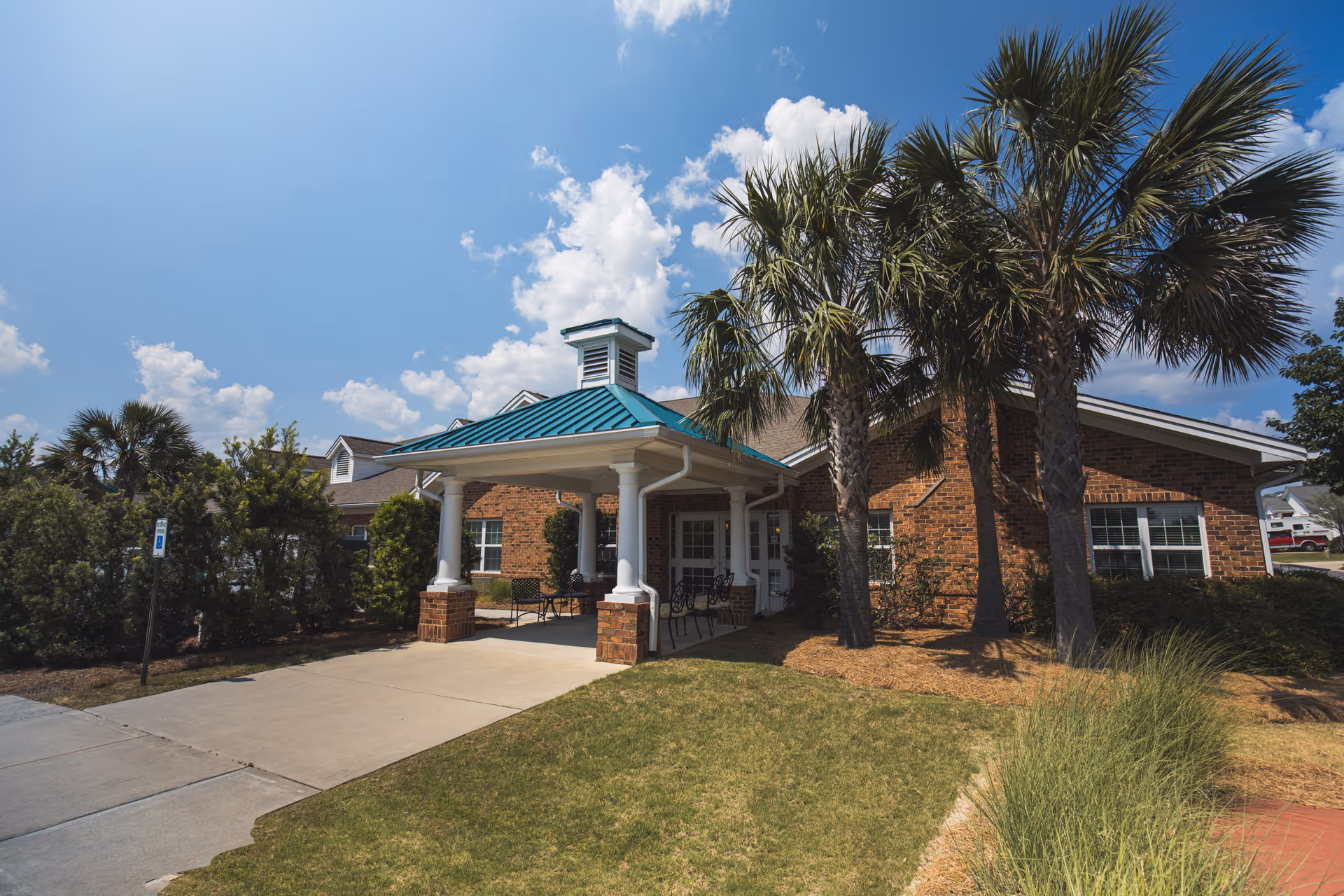 Front entrance of a brick senior living building with a covered portico, palm trees, and a driveway under a blue sky.