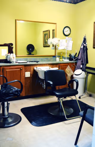 Interior of a small hair salon or grooming area with two black salon chairs, a large mirror mounted on a yellow wall, a countertop with various hair care products, a wall clock, and a white trash bin in the corner.