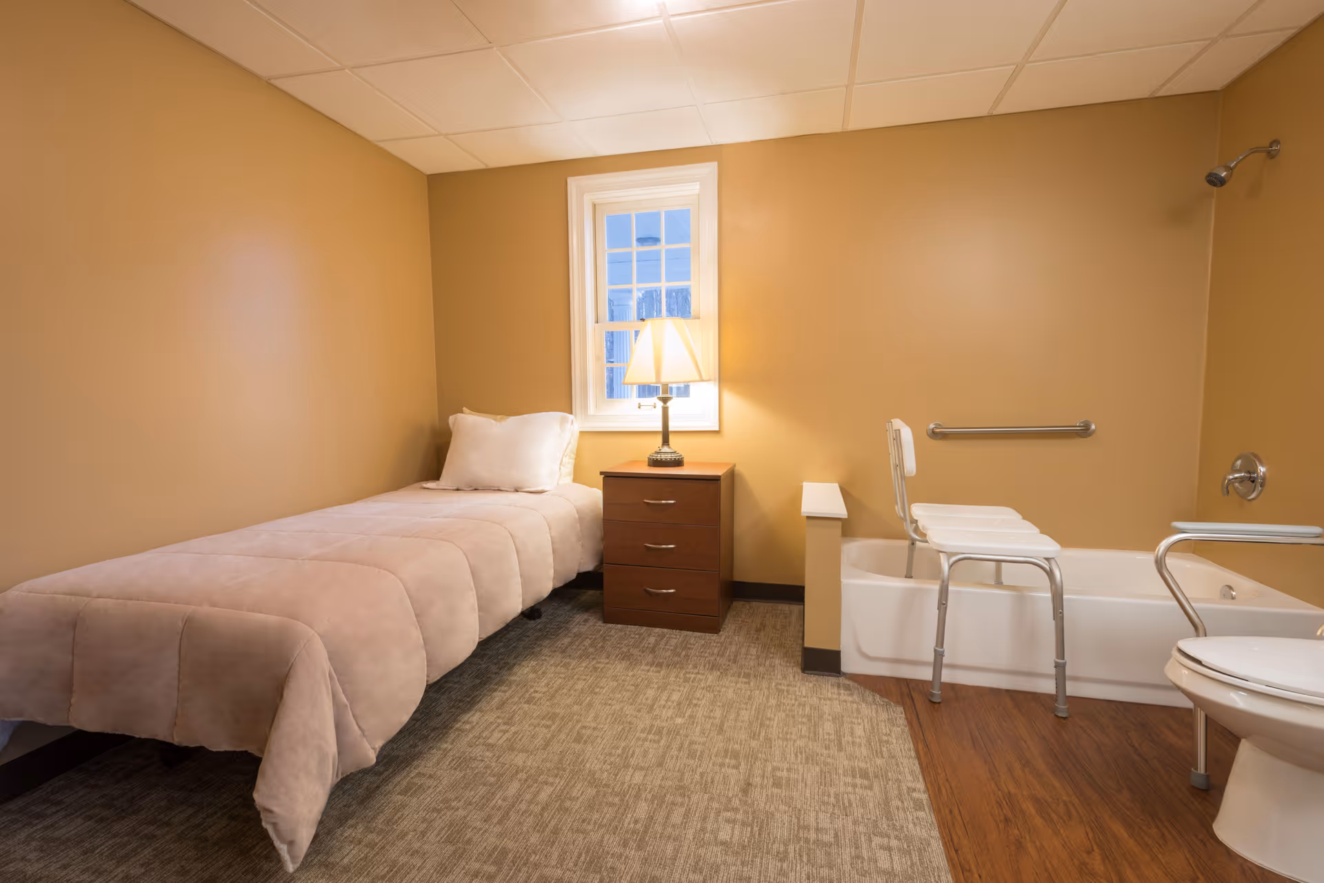 A small room with a single bed covered in a beige comforter, a wooden nightstand with a lamp, a window, and a bathroom area with a bathtub equipped with a shower chair and grab bars, and a toilet with support rails.