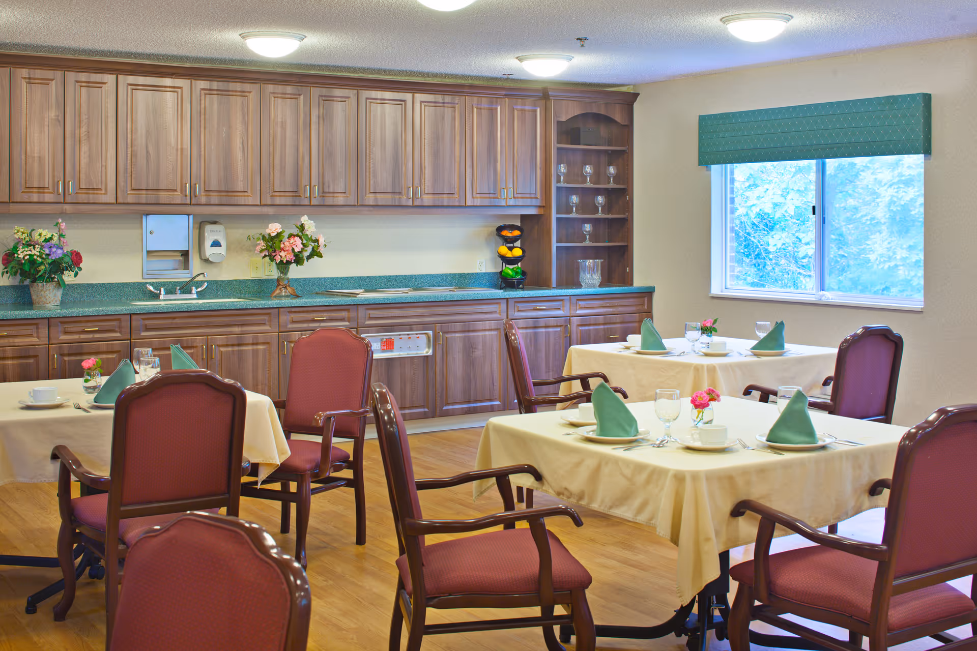 A dining room with four tables covered in beige tablecloths, each set with plates, cups, glasses, silverware, and green folded napkins. The room has wooden chairs with red upholstery, wooden cabinets along the back wall, a countertop with flowers and a fruit stand, and a window with a green valance letting in natural light.