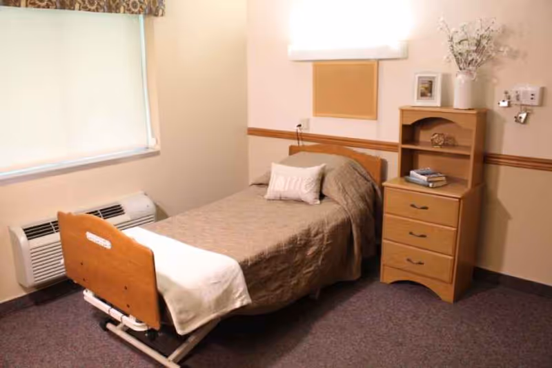 A small, simple bedroom in a senior living facility with a single bed covered in a brown blanket and a white pillow with the word 'Home' on it. Next to the bed is a wooden nightstand with three drawers and an open shelf holding books and a decorative item. A vase with flowers and a framed picture are on top of the nightstand. The room has beige walls with a wooden chair rail, a window with a closed blind, and a wall-mounted air conditioning unit below the window.