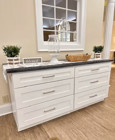 A white sideboard with six drawers topped with a black countertop. On the countertop, there are two small potted plants, a glass water dispenser on a white stand, a woven basket, and a small sign that says 'blessed'. Above the sideboard is a white-framed window with multiple panes reflecting the interior of the room. The floor is wood, and the walls are painted beige.