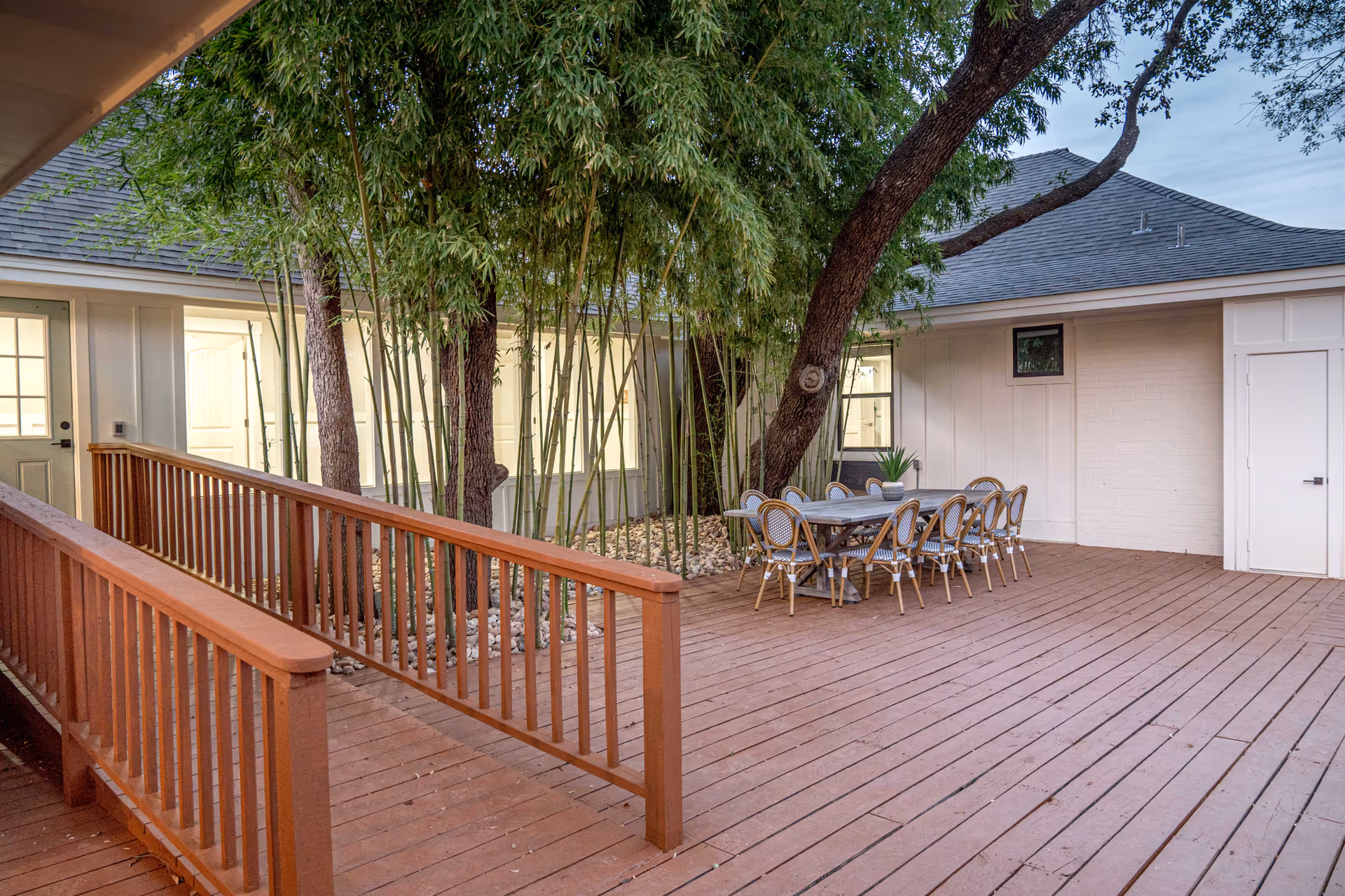 Outdoor wooden deck area with a ramp railing, a dining table with eight chairs, surrounded by trees and plants, adjacent to a building with windows and doors.