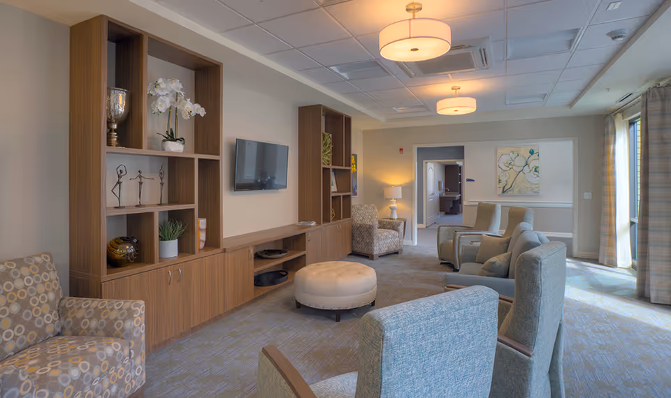 A well-lit living room area in a senior living facility featuring multiple upholstered armchairs arranged around a central round ottoman. The room has built-in wooden shelving with decorative items, a wall-mounted flat-screen TV, and large windows with curtains allowing natural light to enter. The walls are adorned with framed artwork, and ceiling lights provide additional illumination.