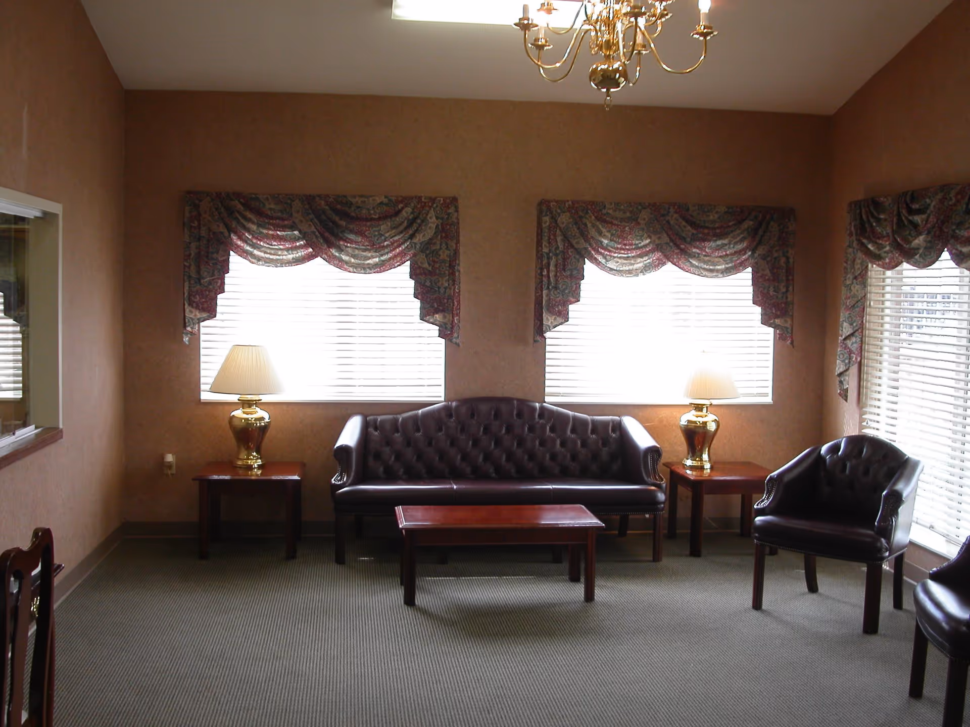 A sitting area with a dark brown leather tufted sofa and two matching armchairs. There are two wooden side tables with gold lamps on either side of the sofa and a wooden coffee table in front. The room has beige walls, carpeted floor, three windows with white blinds and patterned valances, and a brass chandelier hanging from the ceiling.
