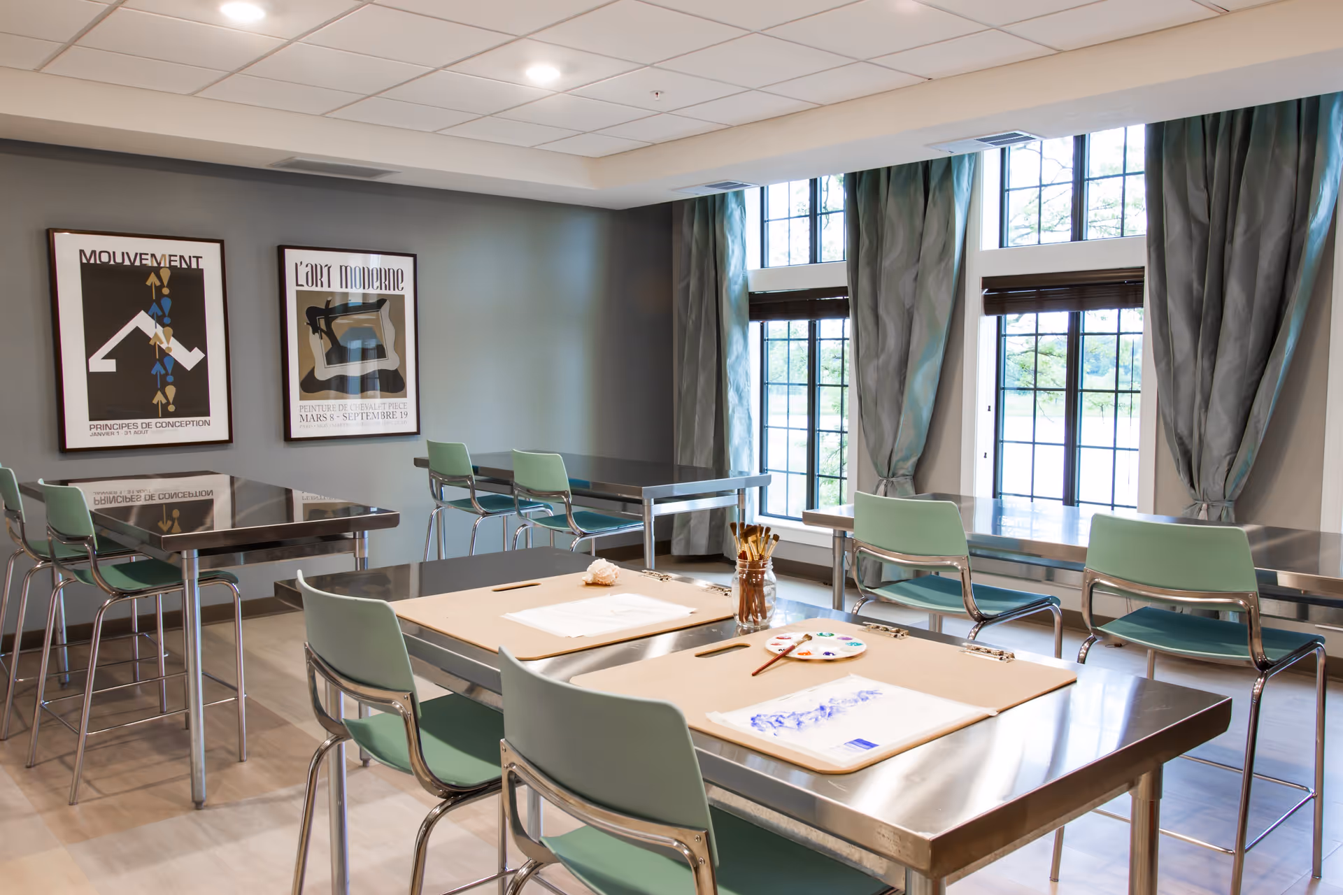 Bright communal activity/dining room with metal tables, mint-green chairs, art supplies on a table, and large windows with curtains.