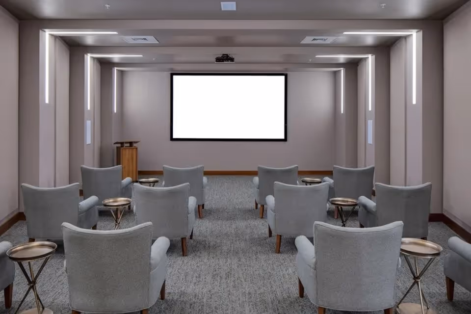 A small theater or presentation room with two rows of gray upholstered armchairs, each paired with a small round side table. At the front of the room is a large blank projection screen mounted on the wall and a wooden podium to the left. The room has modern vertical light fixtures on the walls and ceiling, and a gray carpeted floor.