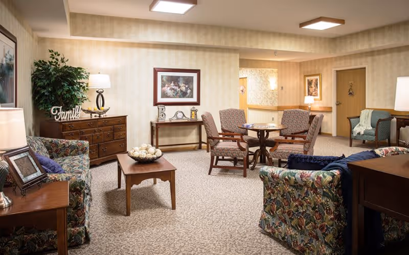 A cozy living room area in a senior living facility featuring floral patterned sofas, wooden tables, armchairs around a small round table, lamps, framed artwork on the walls, and decorative items including a 'Family' sign and a bowl of decorative balls on the coffee table.