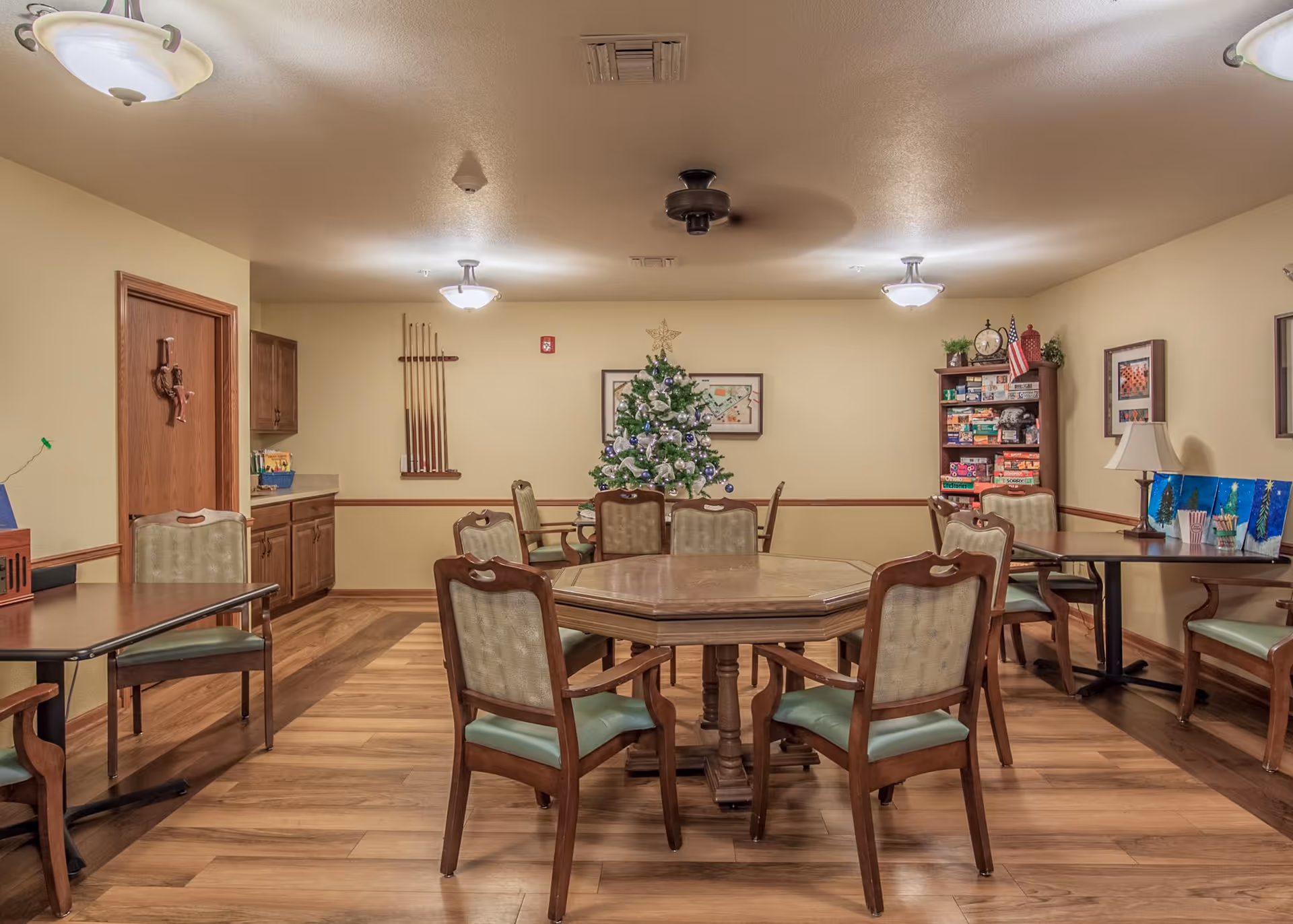 A well-lit common room with wooden floors and beige walls featuring several tables and chairs arranged for group activities. A decorated Christmas tree stands against the back wall, flanked by a rack of board games and framed artwork. The room has ceiling lights and a ceiling fan, with a wooden door and cabinets on the left side.