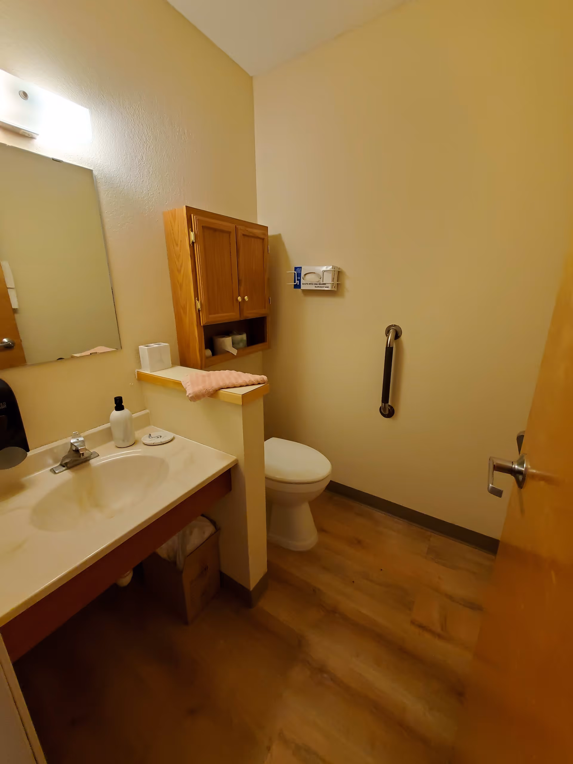 A small bathroom with a wooden floor, beige walls, and a white toilet. There is a sink with a countertop on the left side, a mirror above the sink, a wooden cabinet mounted on the wall above the toilet, and a grab bar on the wall next to the toilet. A pink cloth is placed on the half wall next to the sink.