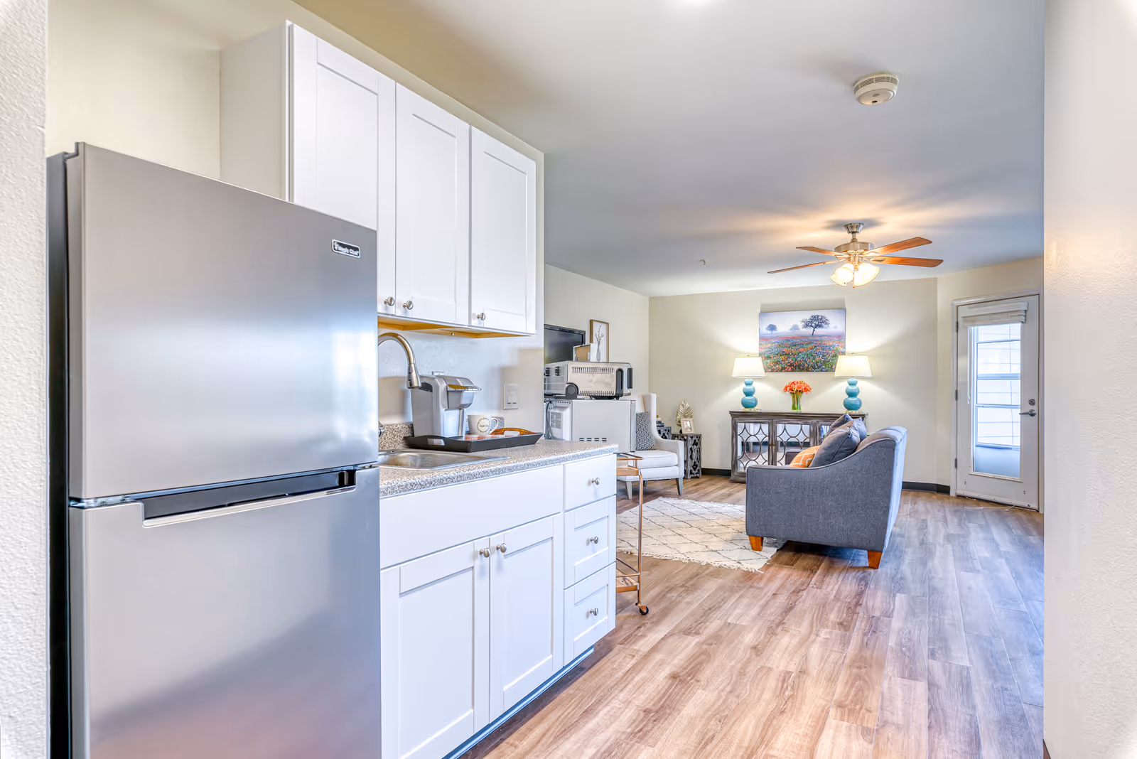 A bright and modern senior living apartment interior featuring a kitchenette with white cabinets, a stainless steel refrigerator, and a coffee maker. The living area includes a gray sofa, an armchair, a patterned rug, a wooden sideboard with two blue lamps and a flower vase, a ceiling fan with lights, and a door leading outside.