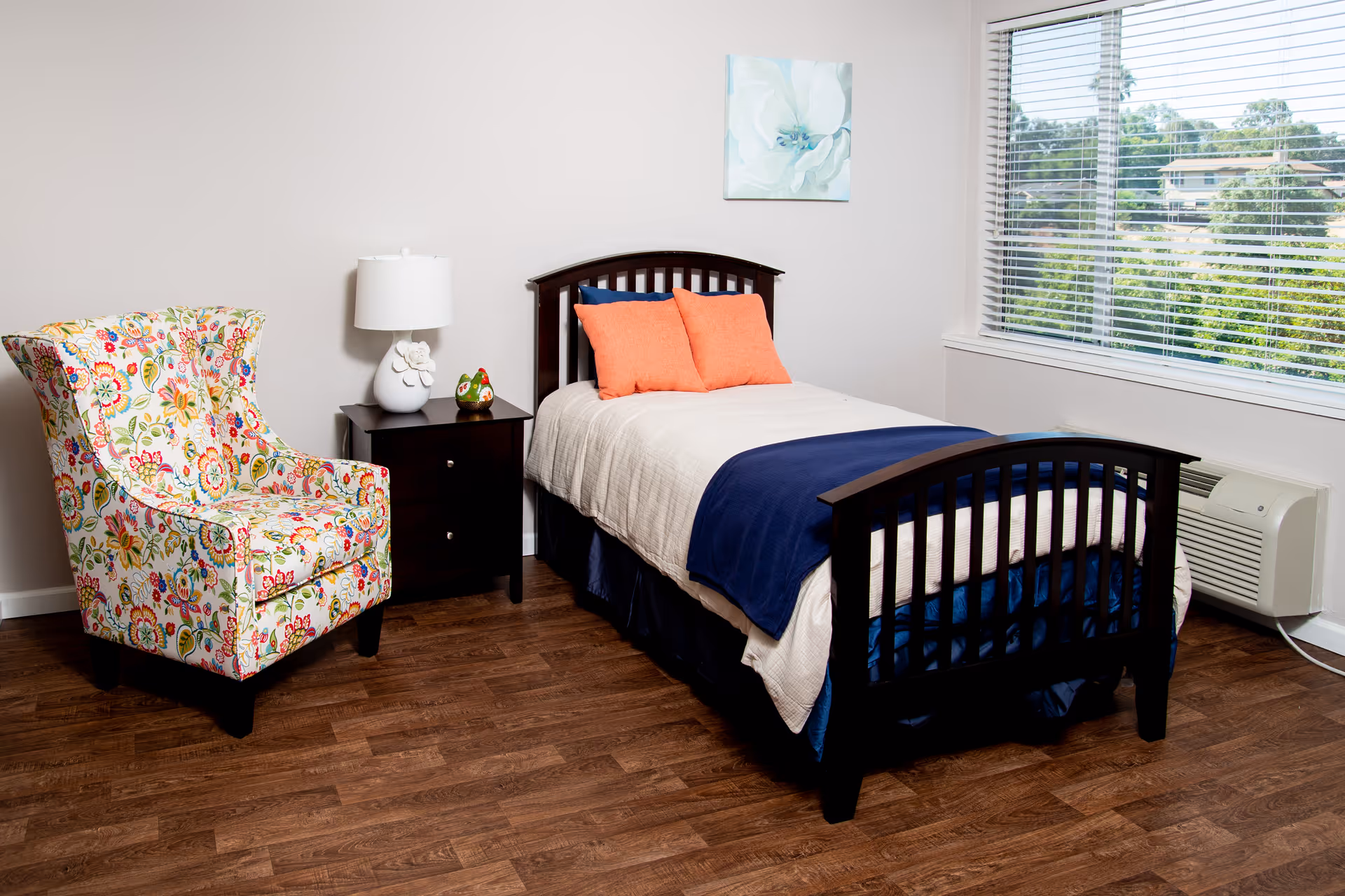 A bright single bedroom with a wooden bed dressed in blue and white linens, a floral armchair, a nightstand with a lamp and small decor, and a window with blinds.