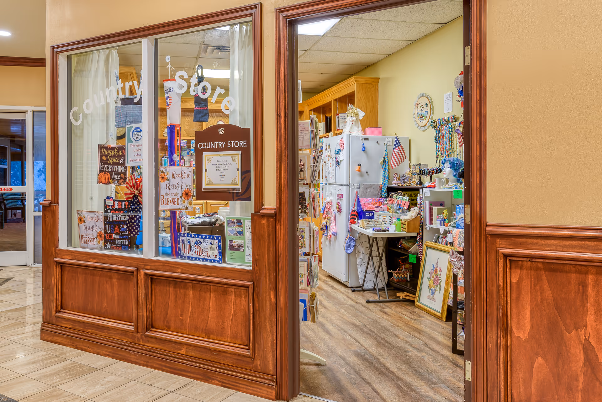 View of a country store inside a senior living facility with wooden paneling and glass windows displaying various decorative items and signs. Inside the store, shelves and a refrigerator are stocked with small goods, crafts, and patriotic decorations.
