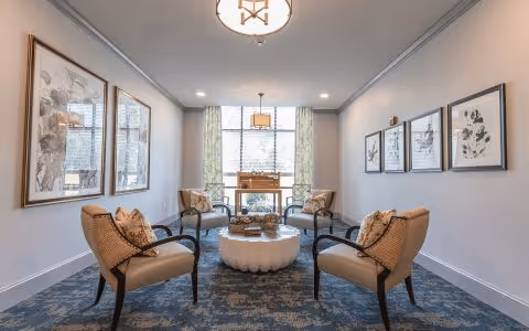 A cozy sitting area with six cushioned armchairs arranged in a circle around a round white coffee table. The room has a blue patterned carpet, light gray walls adorned with framed botanical artwork, and a large window with green curtains letting in natural light. A small table with decorative items is positioned in front of the window.