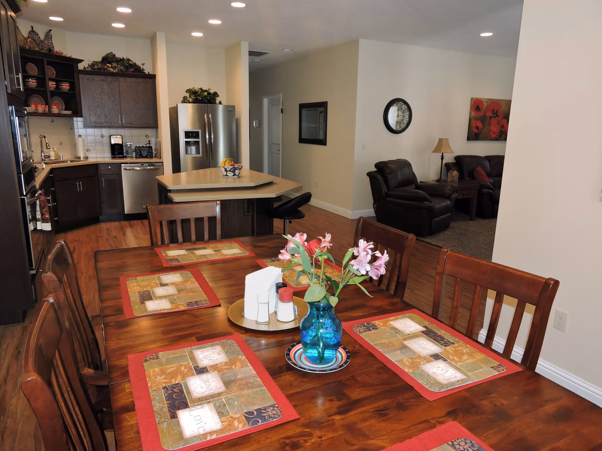 Dining table with placemats and a vase in an open-plan dining area that opens to a kitchen and living room.