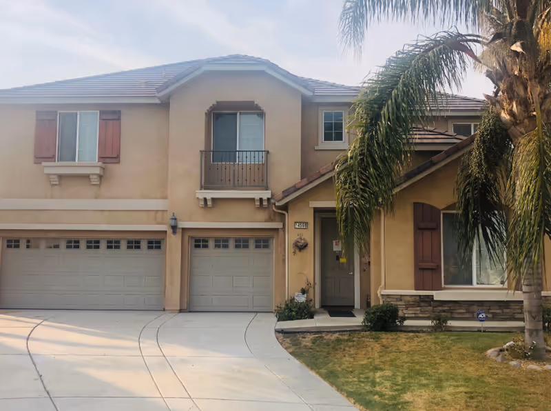 Front exterior view of a two-story beige house with a three-car garage, a small balcony above the middle garage door, and a front door partially obscured by a palm tree. The house has brown shutters and a well-maintained lawn with some shrubs.