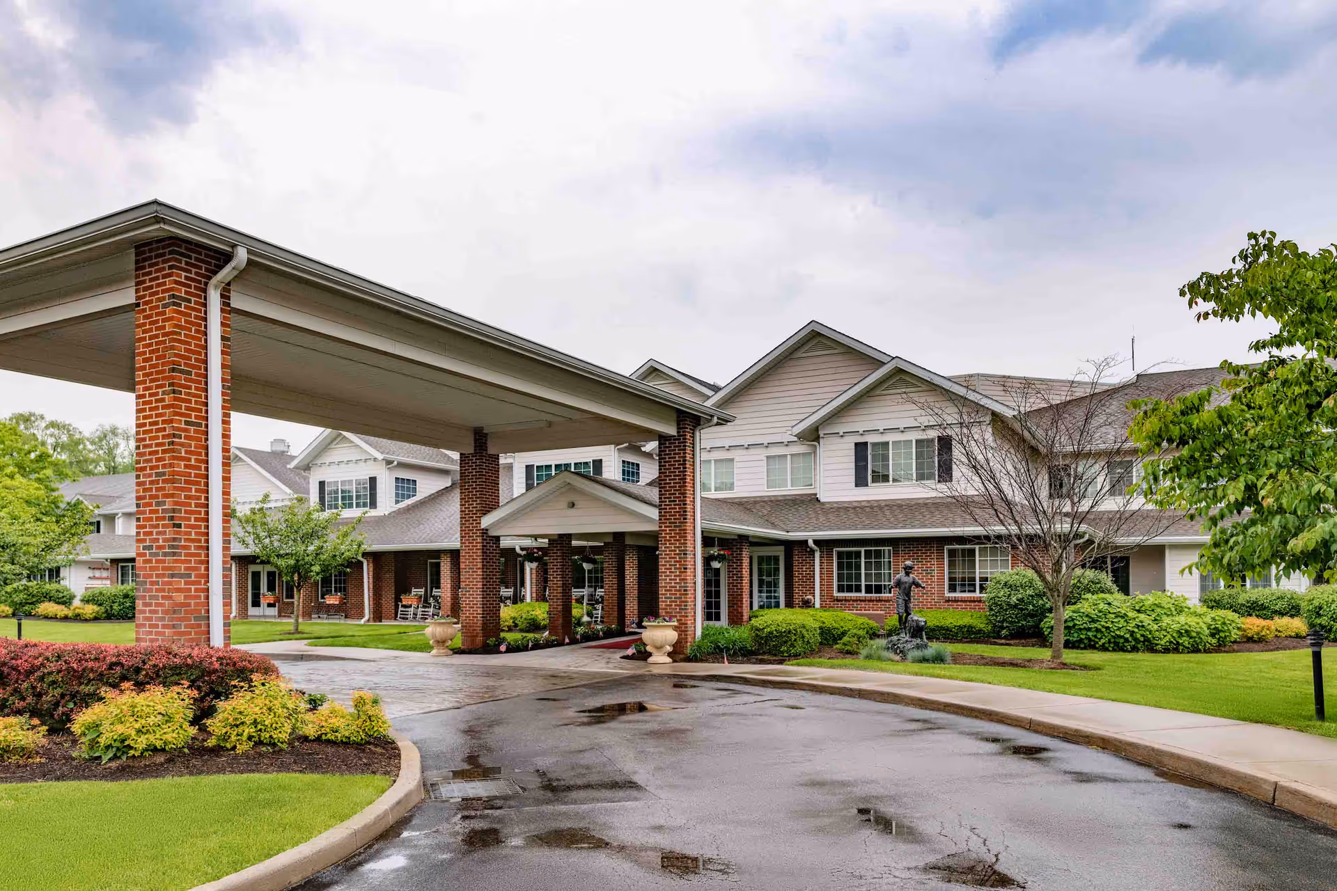 Exterior view of a senior living facility named Holiday Fleming Point showing a covered entrance with brick pillars, a driveway with wet pavement, manicured bushes, trees, and a multi-story building with white siding and brick accents under a cloudy sky.