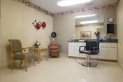 A small room with beige walls and floor, featuring a salon-style chair in front of a large mirror and countertop with various hair care items. There are two chairs and a small table with a plant on the left side, and decorative wall hangings above them.
