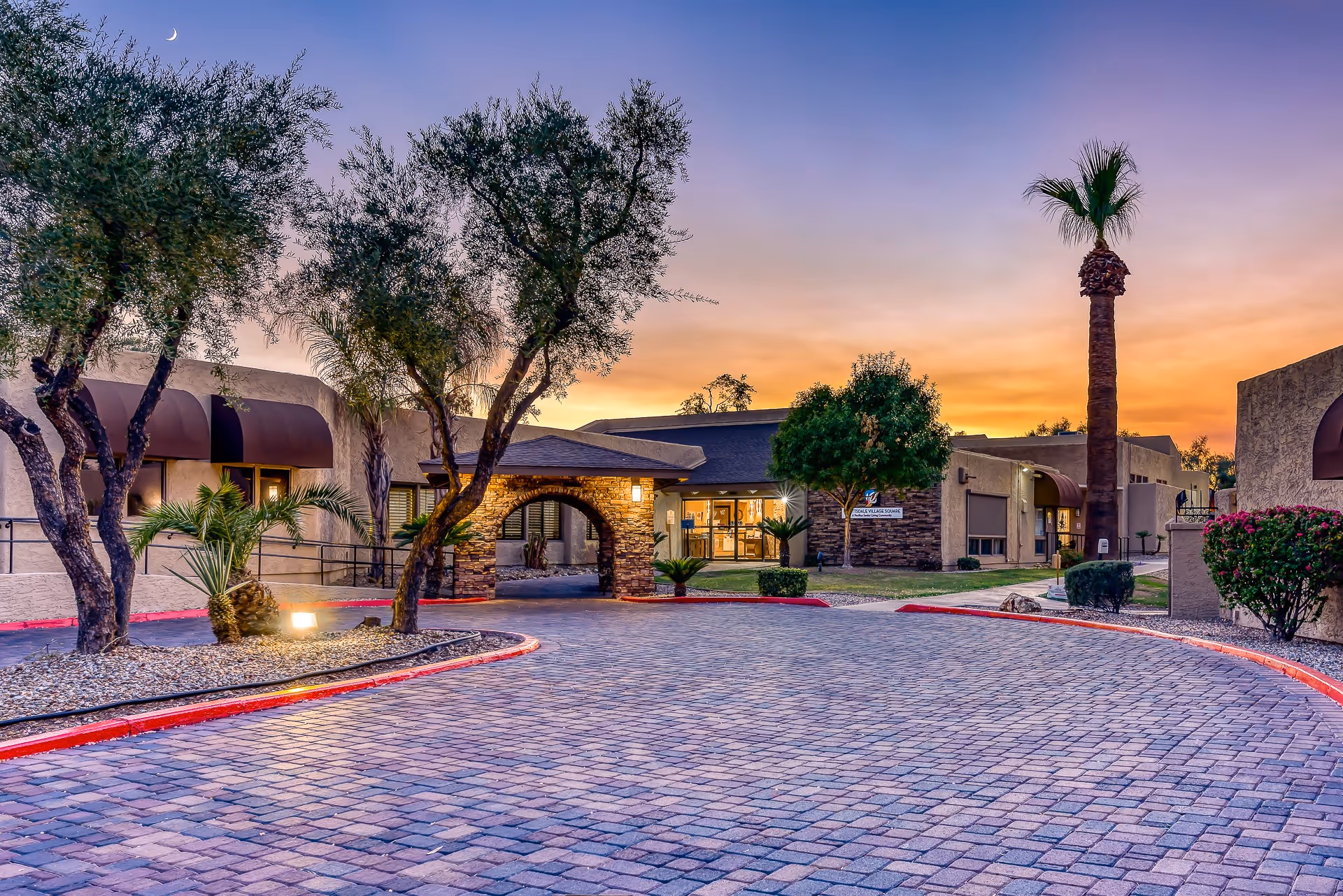 Front entrance of Scottsdale Village Square senior living community at sunset with an arched driveway, palm trees, and a paved courtyard.
