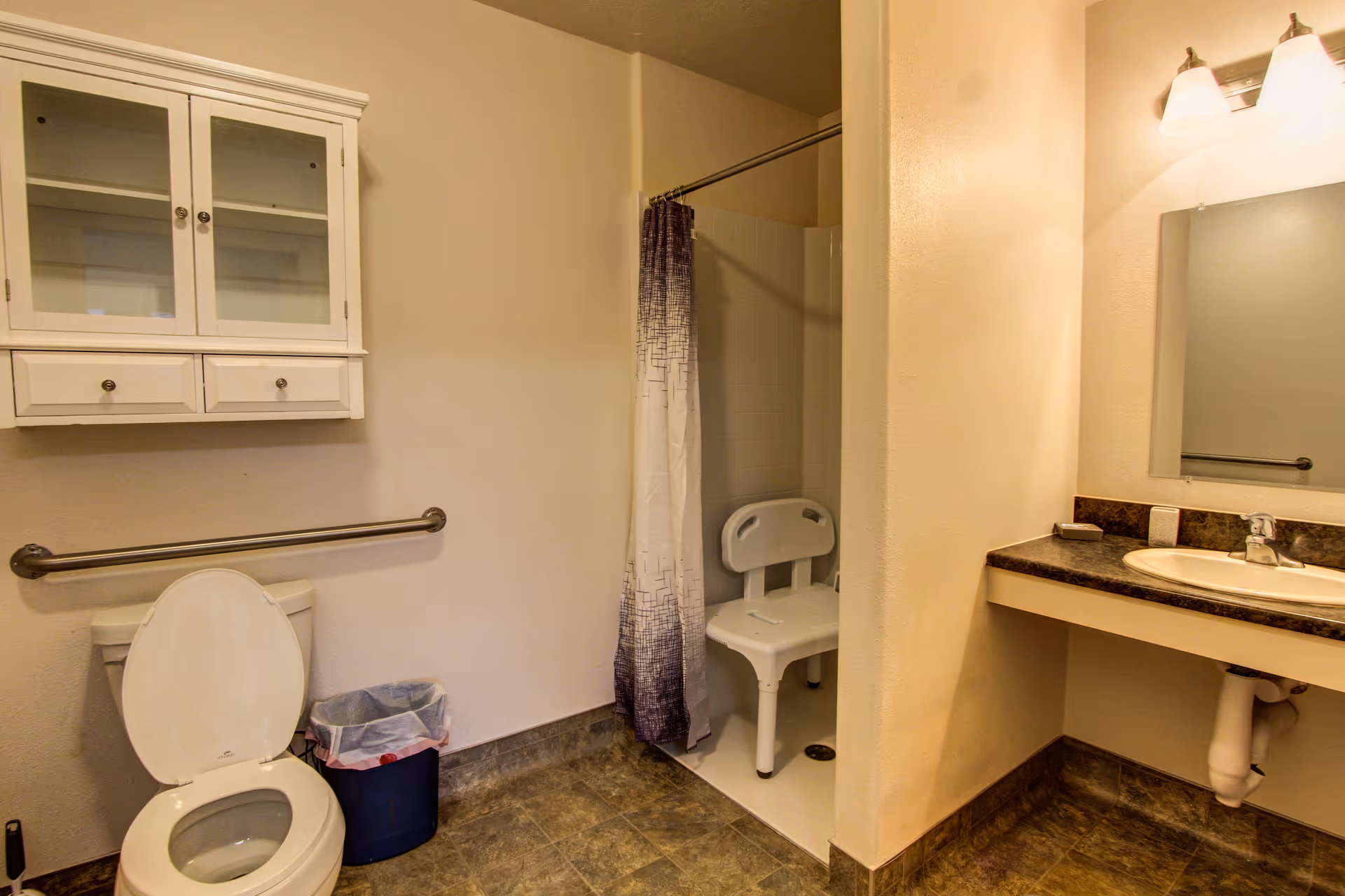 Accessible bathroom with a toilet and grab bar, wall cabinet, walk-in shower with a shower chair, and a sink with mirror.