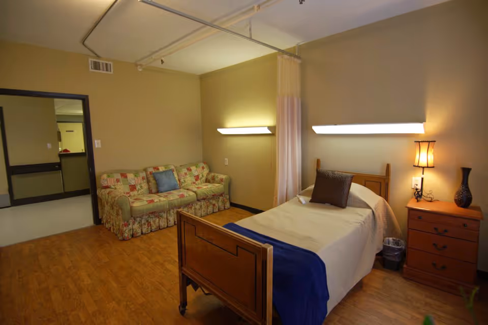 A nursing facility room with a single wooden bed covered with beige bedding and a blue throw at the foot. There is a brown pillow on the bed. Next to the bed is a wooden nightstand with a lamp and a decorative vase. On the left side of the room, there is a floral patterned couch with a blue pillow. The walls are beige, and there are two wall-mounted lights above the bed and couch. The floor is wooden, and there is a curtain hanging from a ceiling track near the bed. An open doorway leads to a hallway outside the room.