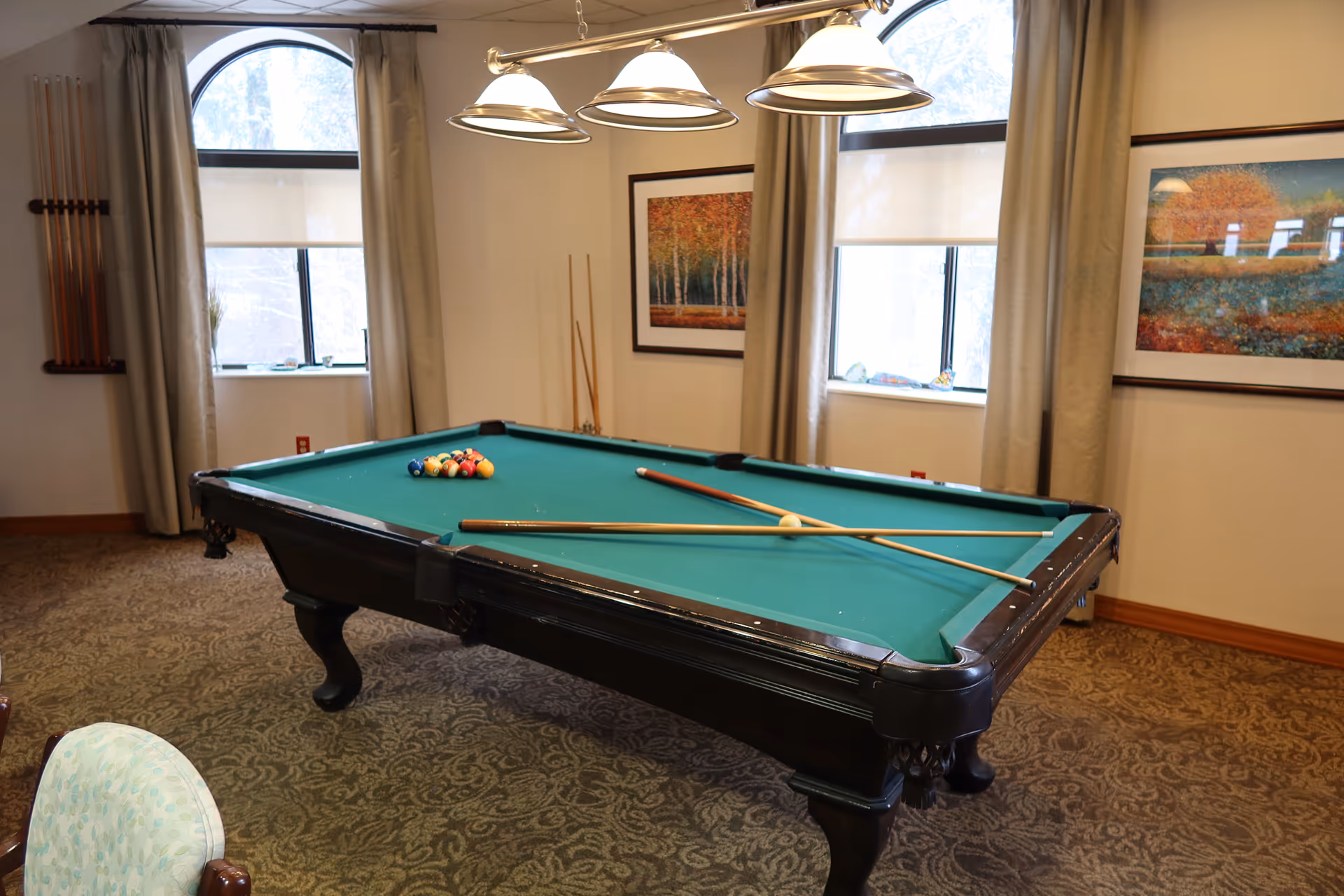 A pool table with cues and racked balls in a well-lit recreation room with windows and framed artwork.