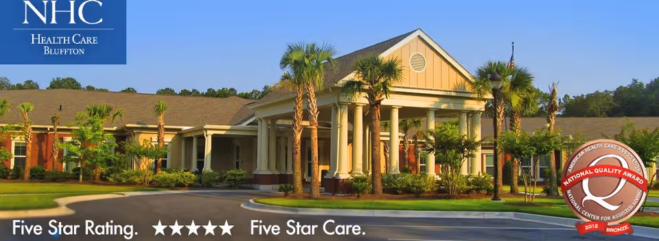 Front entrance of NHC Health Care Bluffton with columns, palm trees, a circular driveway, and an award badge in the corner.