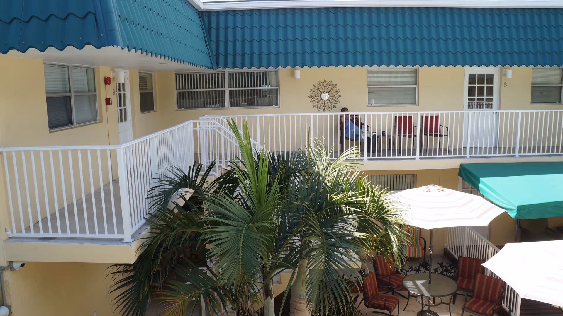 Courtyard with palm trees, patio tables and umbrellas below a second-floor balcony with doors and seating under green awnings.